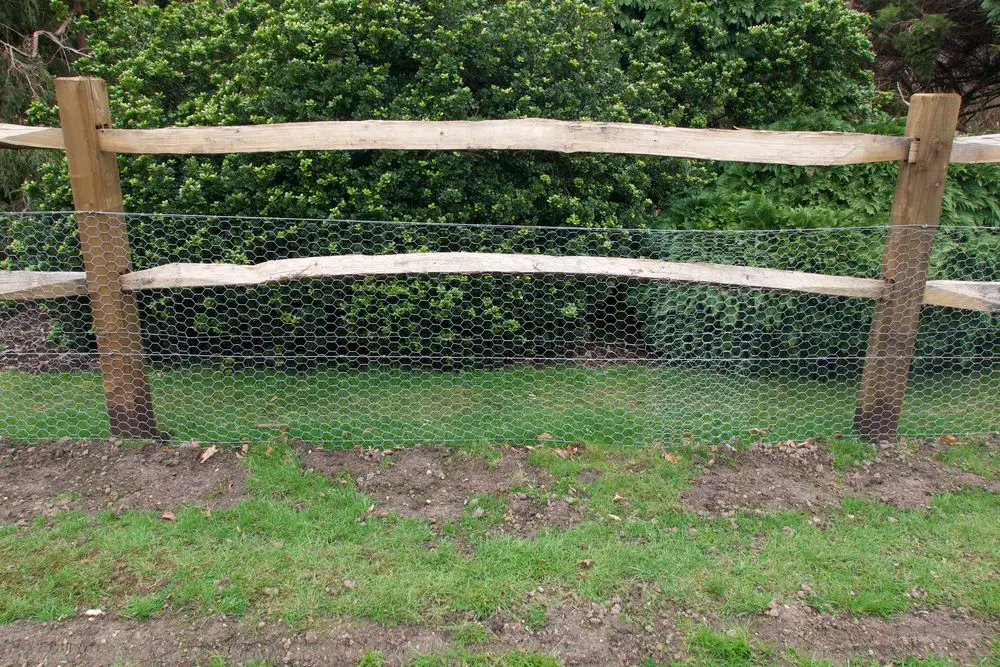 a fence with cattle beams and chicken wire with bushes behind